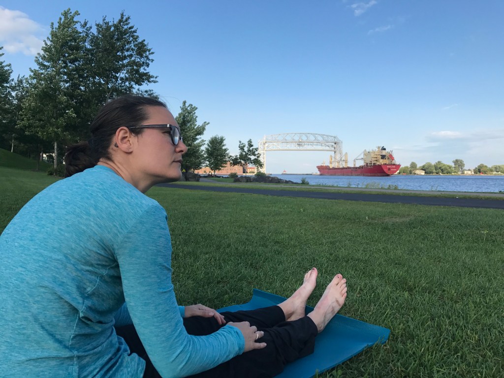 Dr Kristine Snyder, sitting on a mat on a grassy area legs outstretched, barefoot 